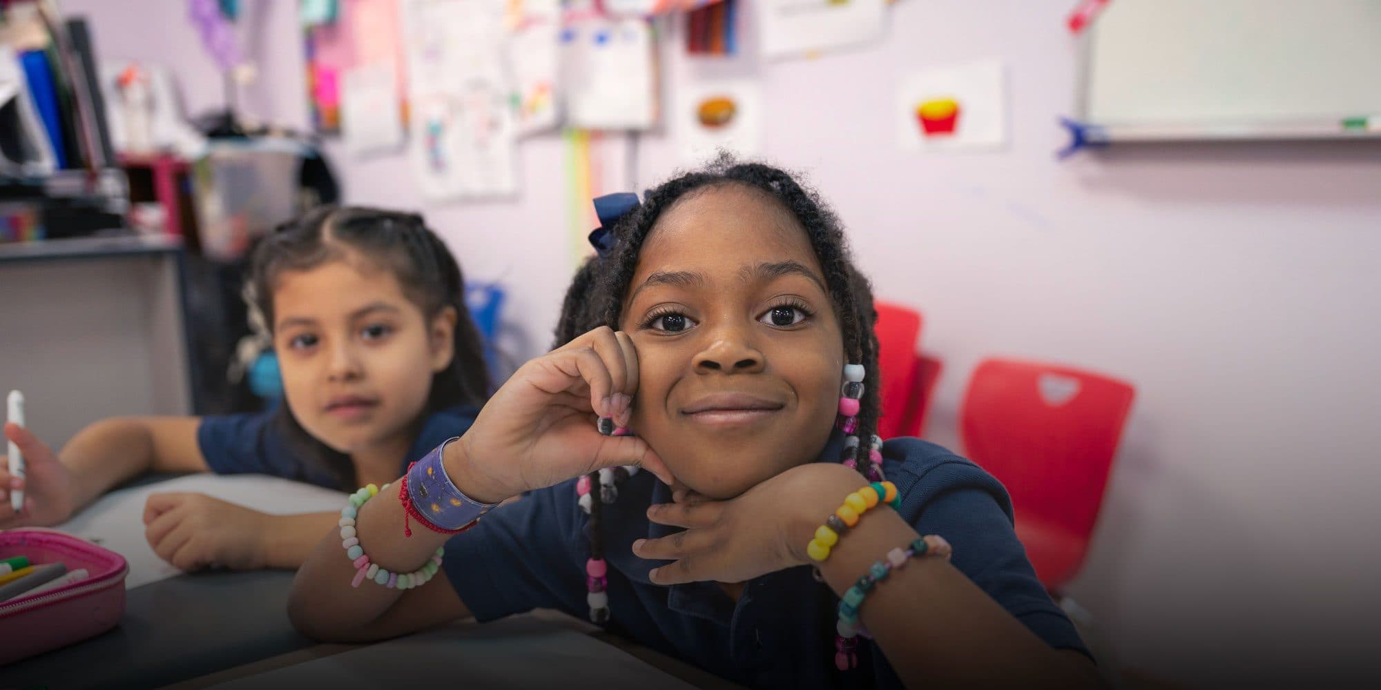 Two young students sit at a classroom table, one smiling with their chin in their hand.