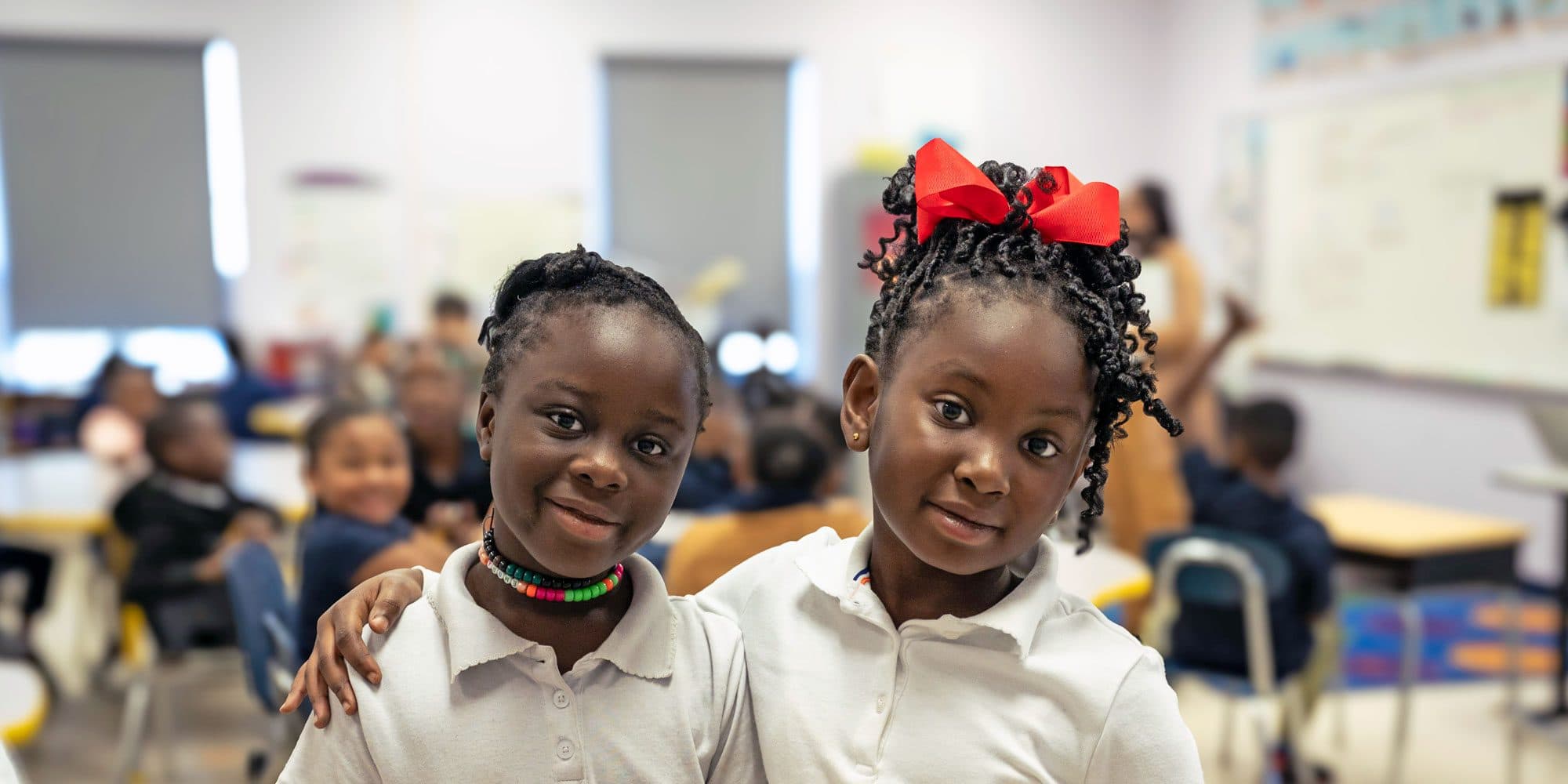 Two smiling students stand arm in arm in a classroom.