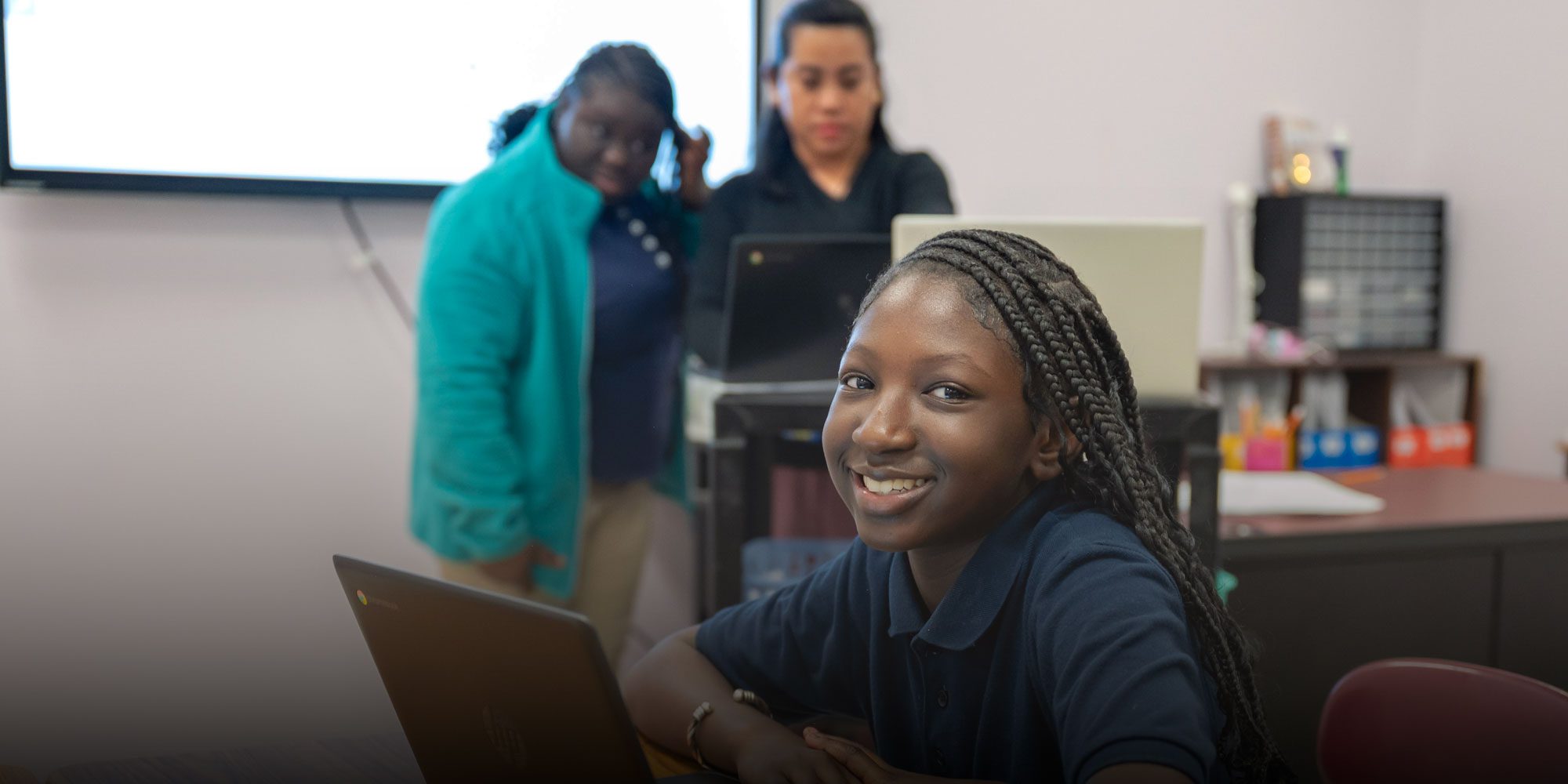 Smiling student with braided hair sits at a laptop in a CHATA classroom.