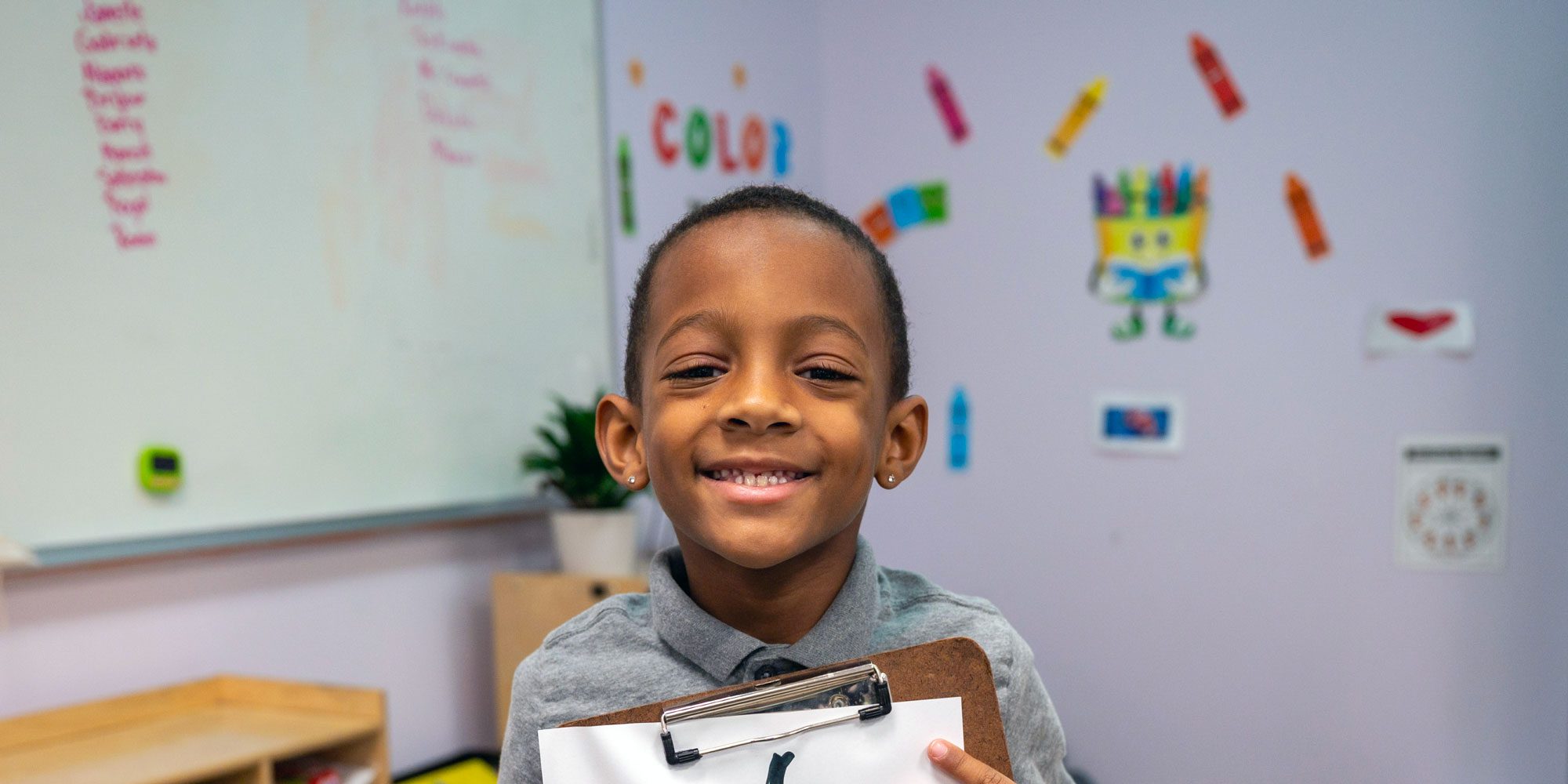 Smiling student holds a clipboard in a colorful classroom.
