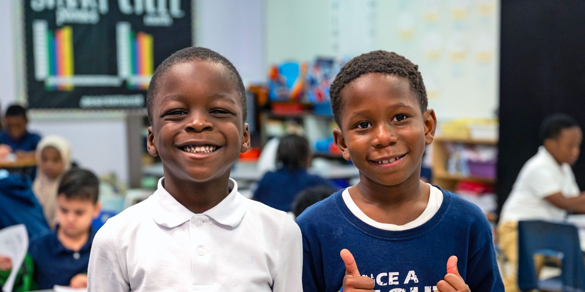 Two smiling boys in a classroom, one giving a thumbs-up.